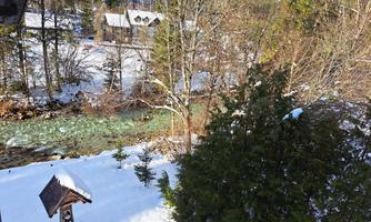 A snowy landscape with a clear river and trees in the foreground. In the background, several huts can be seen.