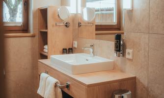 A modern bathroom with wooden furniture, a rectangular sink, and a large mirror. The walls are brightly tiled, and the room has large windows that let in natural light.