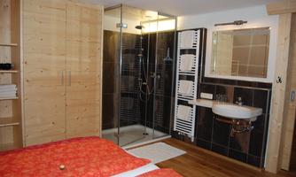 A modern bathroom with a walk-in shower and a sink. In the background, there is a bed with orange bedding and wooden cabinets.