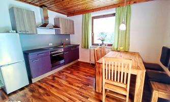 A modern kitchen with wooden components and a dining table for four people.  
Bright windows and green curtains create a friendly atmosphere.