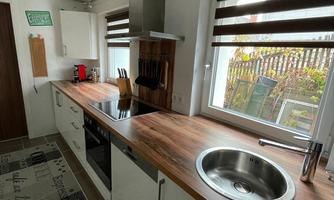 A modern kitchen with a wooden countertop and stainless steel sink. Through the windows, there is plenty of light and a view of the outdoors visible.
