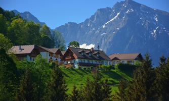A picturesque mountain village with traditional cottages, surrounded by green meadows and dense forests. In the background, majestic mountain peaks rise into the clear blue sky.