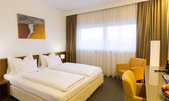 A modern hotel room with two single beds and a desk. Large windows let in plenty of daylight.