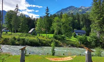 A peaceful landscape with a river, surrounded by trees and mountains. On a meadow, a hammock is hanging between two tree trunks.