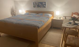 A cozy bedroom with a large double bed and blue bedcovers. Next to the bed are bedside lamps and a small table with a chair.