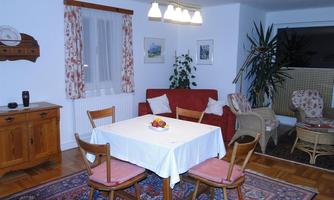 A cozy living area with a table and chairs in the middle. In the background, plants and a comfortable seating area can be seen.