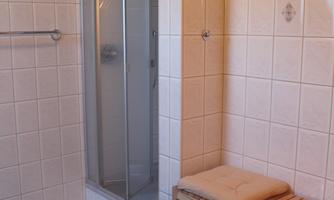 A modern bathroom with a glass shower enclosure. Next to the shower is a wooden shelf with towels and a round bath mat.