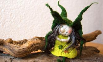 A small figure of a gnome with a green hat and a yellow dress. She is sitting on a piece of wood in front of a bright wall.