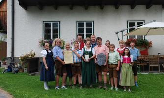 A group of people is wearing traditional Bavarian clothing in front of a house. They are standing together and smiling, while some of them are holding awards in their hands.