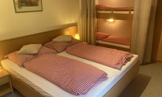 A cozy bedroom with a double bed and a bunk bed in the background. The walls are light, and the bedding is checked.