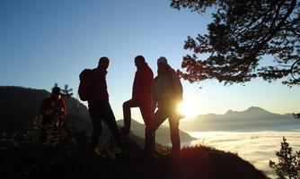 A group of hikers stands on a hill at sunrise. The sky is clear and the sun shines over a blanket of clouds.