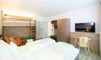 A modern hotel room with two single beds, a loft bed, and a television. There is also a small desk and a chair.