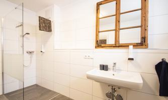 A modern bathroom with a rain shower and an elegant sink. Generous tiles and a wooden window create a bright, inviting atmosphere.
