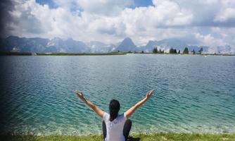 A person is sitting by the shore of a clear lake, spreading their arms. In the background, mountains and a cloudy but bright sky can be seen.