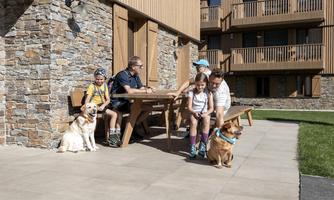 A family is sitting outside at a table with two dogs. In the background, modern hotel buildings and a green meadow can be seen.