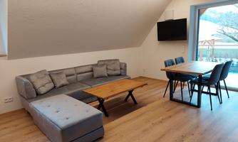 A modern living room with a comfortable gray sofa and a wooden table. The room has large windows that let in plenty of light.