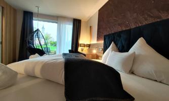 A modern bedroom with a cozy bed and elegant decor. In the background, there is a window overlooking greenery and a stylish hanging chair.