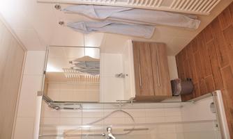 A modern bathroom with a shower, a sink, and a wooden shelf. In the background, two grey bathrobes are hanging.