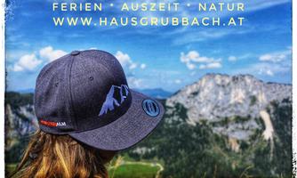 A person with a grey cap is standing in front of a picturesque mountain landscape. The view shows high mountains under a blue sky.