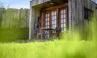 A small wooden house with a terrace and two chairs. In front of the house lies lush, green grass.