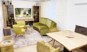 A modern living room with a cozy green sofa and a wooden table. A television hangs on the wall, and there are shelves with books.