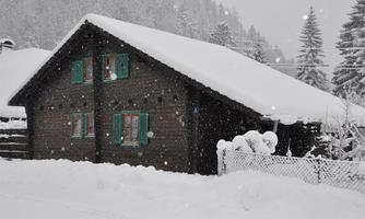 A cozy wooden house in a snowy landscape. The snow covers the roof and the surroundings, while small spruces are visible in the background.