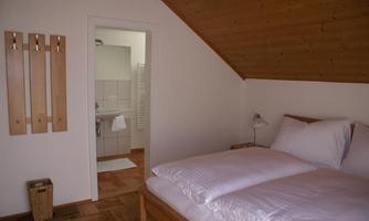 A cozy bedroom with a double bed and wood paneling. In the background, there is a bathroom with light tiles.