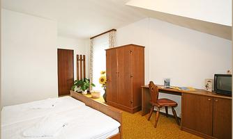 A cozy bedroom with two beds and a wooden wardrobe. A desk and a chair complement the furnishings in a bright room.
