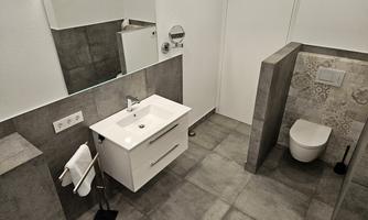 A modern bathroom with a white sink unit and a separate toilet. The walls are in neutral gray tones and have decorative tiles.