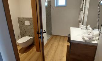A modern bathroom with wooden floors and bright walls. There is a shower, a sink, and a toilet, as well as towels on the shelf.