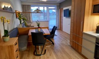 A modern living room with a wooden dining area and a cozy seating corner. Large windows let in plenty of light into the room.