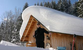 A man stands in front of a wooden cabin surrounded by snow. A thick layer of snow lies on the roof under a clear blue sky.