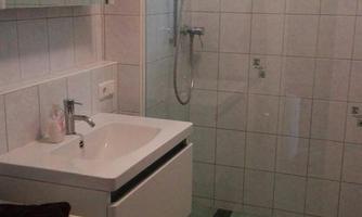 A modern bathroom with a shower, a sink, and a washing machine. The walls are tiled and there is a window for natural light.