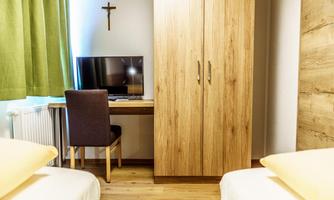 A simple room with two beds, a wooden wardrobe, and a desk. A television is on the desk, and a cross hangs on the wall.