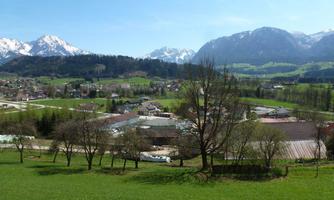 A picturesque landscape with green meadows, a small village, and snow-capped mountains in the background. The sky is clear and blue, creating a peaceful atmosphere.