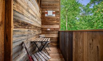 A small balcony with wood cladding and a table with two chairs. In the background, green trees are visible.
