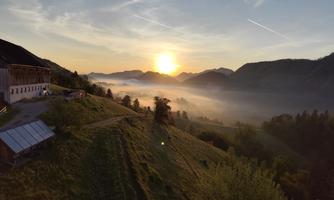 A picturesque sunrise over a hilly landscape with gentle wisps of fog. In the foreground, there is a farmhouse surrounded by green grass and trees.