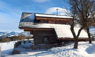 A cozy wooden house in the snow with a snow-covered roof. In the background, gentle hills and a clear blue sky can be seen.