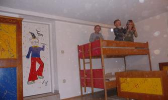 Three children are standing on a loft bed in a colorful room. On the wall, you can see a funny picture of a clown.