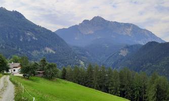 A picturesque mountain landscape with gentle hills and dense trees. In the foreground, a path leads to a small house in nature.