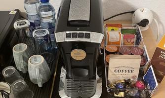 A modern coffee machine sits on a table with glasses and bottles of water. Next to the machine is a selection of coffee capsules and tea bags.