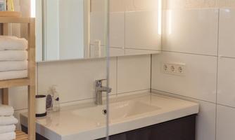 A modern bathroom with a sink and a large mirror. Next to the sink is a shelf with towels.