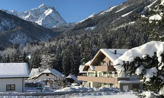 A snow-covered landscape with a modern building and traditional chalets. In the background, a majestic mountain range rises under a clear blue sky.