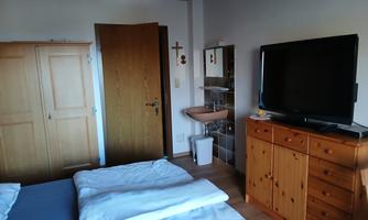 A cozy bedroom with a double bed, a television, and wooden cabinets. The walls are light and the floor is wooden.