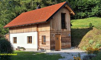 A modern wooden house with a red roof, surrounded by green landscape. The facade combines wood and plaster elements and features large windows.