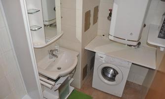 A small bathroom with a sink, a shower, and a washing machine. The walls are brightly tiled and there is a green carpet on the floor.