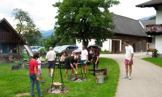A group of people is grilling outdoors on a meadow. In the background, houses and trees can be seen.