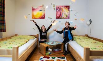 Two children laugh and throw banknotes into the air. The room has two single beds and colorful wall paintings.