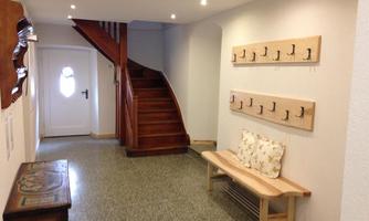A bright hallway with a wooden staircase and a white door. Several coat hooks are hanging on the wall and a bench is available.