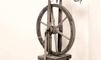 An old wooden spinning wheel stands in the corner of a room. It has a classic design with a large wheel and a seat.